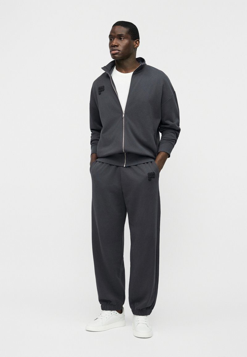 Homme debout les mains dans les poches, portant un survêtement zippé gris foncé et des baskets blanches sur un fond clair uni.