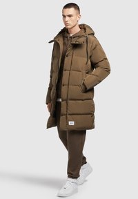 Pruun puffjakken koos kapuutsiga, millel on tikkimisdisain ja esine nupud. Kandmiseks sobiva pruuni kostüümi ja valgete tossudega.