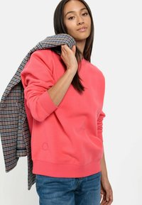 Korallenfarbener Sweatshirt mit gerippten Bündchen und Halsauschnitt, mit einem kleinen Logodetail am Saum. Darüber wird ein karierter Blazer getragen.