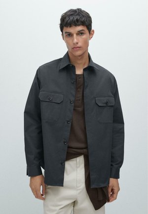 Dark gray button-up jacket with two chest pockets, durable fabric, pointed collar, and long sleeves. Worn over a brown shirt.