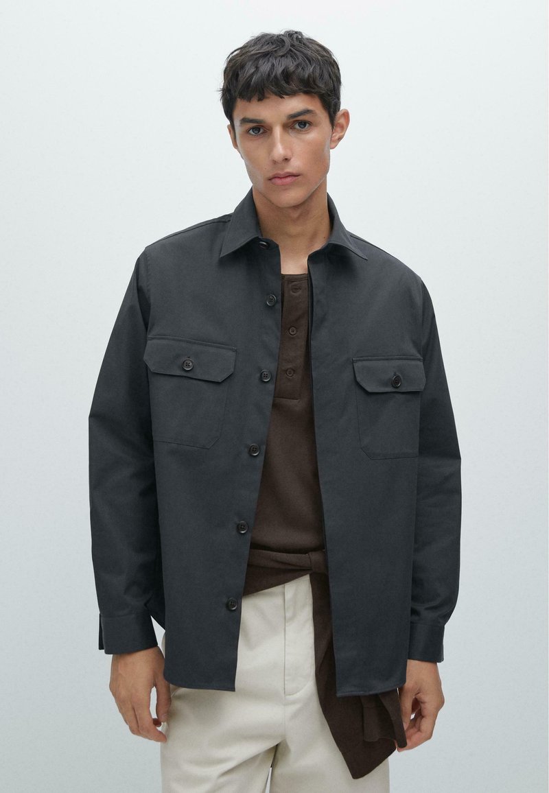 Dark gray button-up jacket with two chest pockets, durable fabric, pointed collar, and long sleeves. Worn over a brown shirt.