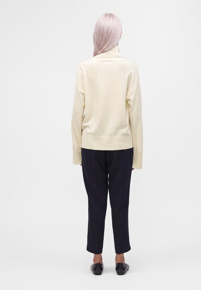 Personne aux longs cheveux pâles portant un pull à col roulé crème, un pantalon noir et des chaussures noires, debout de dos sur un fond uni.