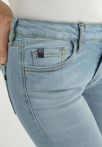 Jean bleu clair avec un design ajusté, deux poches avant, une petite étiquette de marque et des détails de couture jaunes tout au long.