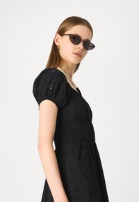 Μαύρο φόρεμα με φουσκωτά μανίκια, λουλουδάτη κεντημένη διακόσμηση, ελαστική μέση και φαρδιά φούστα. Φοριέται με μαύρα γυαλιά και κολιέ με πέρλες.