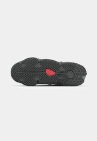 Black shoe sole with a textured grip pattern. A small red emblem is centrally located. The background is plain white.