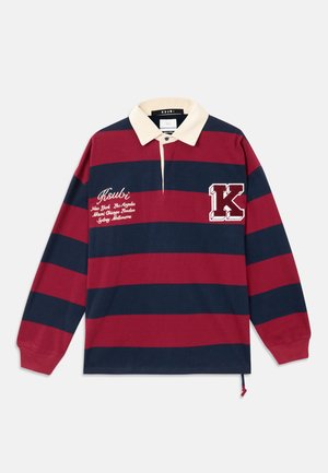 La camiseta polo de manga larga presenta rayas horizontales en rojo y azul marino, un collar beige, un logo bordado y un parche decorativo con la letra "K".