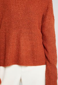 Pull tricoté orange rouille à manches longues avec un motif texturé, associé à un pantalon blanc cassé, visible du torse jusqu'au milieu des cuisses.