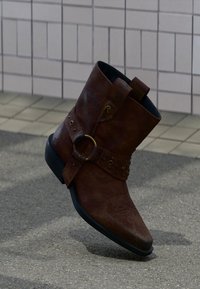 Καφέ δερμάτινο μποτάκι αστραγάλου με διακοσμητικό λουράκι, μεταλλικό δαχτυλίδι και τρουκς, τοποθετημένο σε μια πλακόστρωτη εξωτερική επιφάνεια με πλακάκια και λιθόστρωτο.