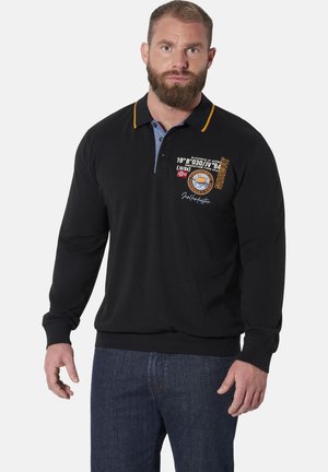 Bearded man wearing a black long-sleeve polo shirt with printed details and a blue button placket, paired with dark blue jeans.