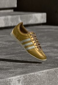 Guldig skinnsneaker med tre vita ränder, bruna snören, perforerade detaljer och en texturerad sula, visad mot en grå bakgrund.