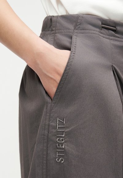 Χέρι τοποθετημένο στην τσέπη γκρι παντελονιού με την επωνυμία Stieglitz, που φοριέται με λευκό τοπ, δείχνοντας την υφή του υφάσματος και τις λεπτομέρειες ραφής.