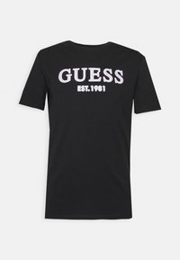 Sort bomulds T-shirt med rund halsudskæring og korte ærmer. Foran vises "GUESS" i hvid og pink tekst, sammen med "EST.1981".
