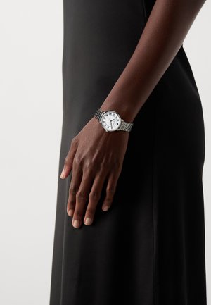 Montre-bracelet argentée avec un bracelet métallique texturé, cadran blanc présentant des chiffres romains et trois aiguilles noires sur un fond noir discret.