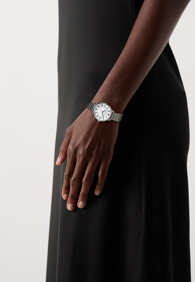 Silverfärgad armbandsur med en texturerad metallarmband, vit urtavla med romerska siffror och tre svarta visare på en dämpad svart bakgrund.