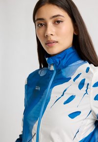 Blå och vit jacka med abstrakta akvareller, mjuk textur, hög krage och kontrasterande blå dragkedja, med en logotyp på framsidan.