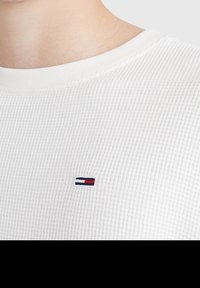 Bílý bavlněný svetr s vaflovou pleteninou a kulatým výstřihem, na hrudi s malým vyšitým logem v námořnické modré, červené a bílé barvě.