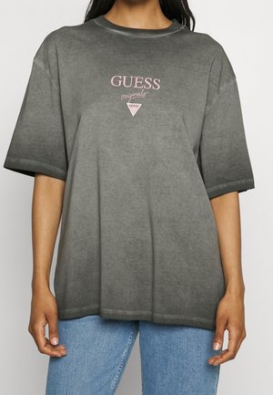 Kvinde iført falmet grå oversized T-shirt med pink "GUESS originals" logo, kombineret med lyseblå jeans, vist fra nakke til hofter.
