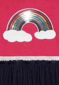 Fuksia kangas, jossa on kimalteleva sateenkaaren kuvio, hopeiset yksityiskohdat ja valkoiset pilvimuotokuvat, yhdistettynä laivastonsiniseen täyshameen alaosaan.