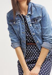 Jeansjacka med en ljusblå tvätt, knappad framtill, bröstfickor och uppkavlade ärmar, bärs över en marinblå prickig klänning med ett brunt bälte.