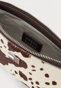 Kott, millel on pruun ja koorevärvi lehmade print, mis sisaldab lukuga taskut, pruuni sisekatte ja sildi "TOMMY JEANS".