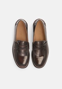 Mocasines de cuero marrón oscuro con un acabado suave, que cuentan con una correa decorativa en la parte superior y costuras en contraste a lo largo de los bordes.
