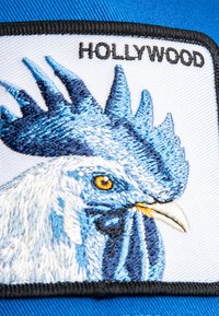 Parche bordado de la cabeza de un gallo azul y blanco con ojo naranja, borde negro y la palabra "HOLLYWOOD" arriba sobre un fondo de tela azul.