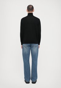 Svart tröja med hög krage, tillverkad av texturerat tyg, i kombination med ljusblå utsvängda denimjeans. Visas från baksidan.