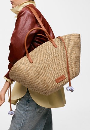 Persona que lleva un gran bolso de mano tejido en tono marrón claro con asas de cuero y un parche de cuero, vistiendo chaqueta de cuero roja y vaqueros azul claro.