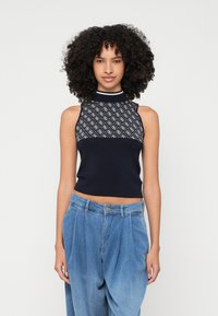 O tânără cu păr creț, îmbrăcată cu un top fără mâneci de culoare bleumarin, cu un model geometric, și cu o pereche de blugi largi, cu talie înaltă, stând în fața unui fundal alb.