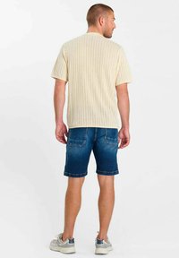Homme de dos portant une chemise à manches courtes texturée crème, un short en denim bleu et des baskets grises sur un fond blanc uni.