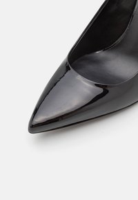 Chaussure à talon haut en cuir verni noir avec un bout pointu et une finition lisse et brillante. Intérieur lisse et arrondi à l'ouverture.