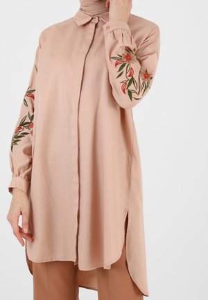 Beige lange mouwen tuniek met bloemenborduursel op de mouwen, voorzijde met knopen, zijsplitten, gedragen met bijpassende broek.