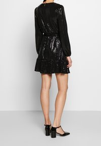 Robe noire à paillettes avec des manches longues, taille cintrée et ourlet à volants superposés. Assortie à des escarpins noirs en vernis avec une lanière.