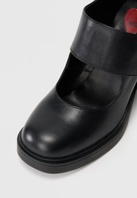 Sapato de couro preto com biqueira arredondada, design recortado e tira larga no tornozelo. Apresenta uma textura suave e sola elevada e robusta.