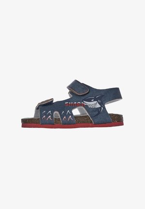 Sandal de denim azul con diseño de tiburón, correa ajustable, suela de corcho, acentos rojos, estilo de punta abierta, cortes para ventilación, detalles de olas juguetonas.