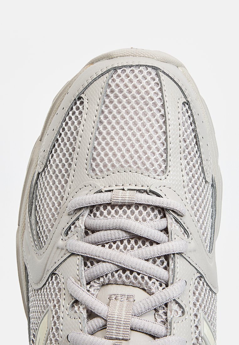 Zapatilla gris claro con parte superior de malla y acentos texturizados, puntera redondeada y cordones planos. Sobrelaminados de plástico que destacan en los laterales.