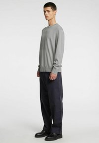 Hall hallaste pikkade varrukatega kampsun ümmarguse kaelusega, pehme tekstuuriga ja soonikkoes varrukate äärtega, kombineeritud lahtiste tumedate pükstega ja mustade kingadega.