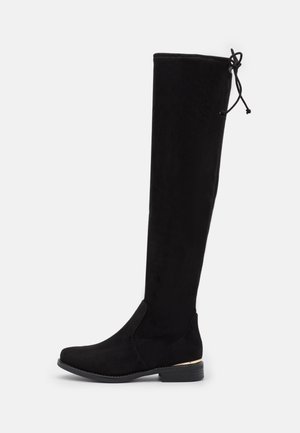 Bottes noires au-dessus du genou, fabriquées en matériau doux semblable à du suède, avec un talon plat, un accent à la cheville et un fin lien en haut pour l'ajustement.
