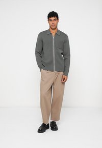 Mann som har på seg en grå zip-up genser, beige brede bukser og svarte sko, stående foran en enkel hvit bakgrunn.