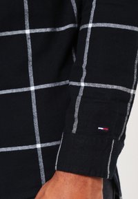 Maglietta nera a maniche lunghe con motivo a scacchi bianchi, maniche con polsino e un piccolo logo di marca rosso, bianco e blu vicino al polso.
