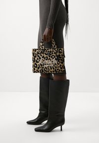 Sac à main à imprimé léopard avec le logo "Coach" brodé, doté de poignées doubles. Le modèle porte une robe moulante à côtes et des bottes noires hautes.
