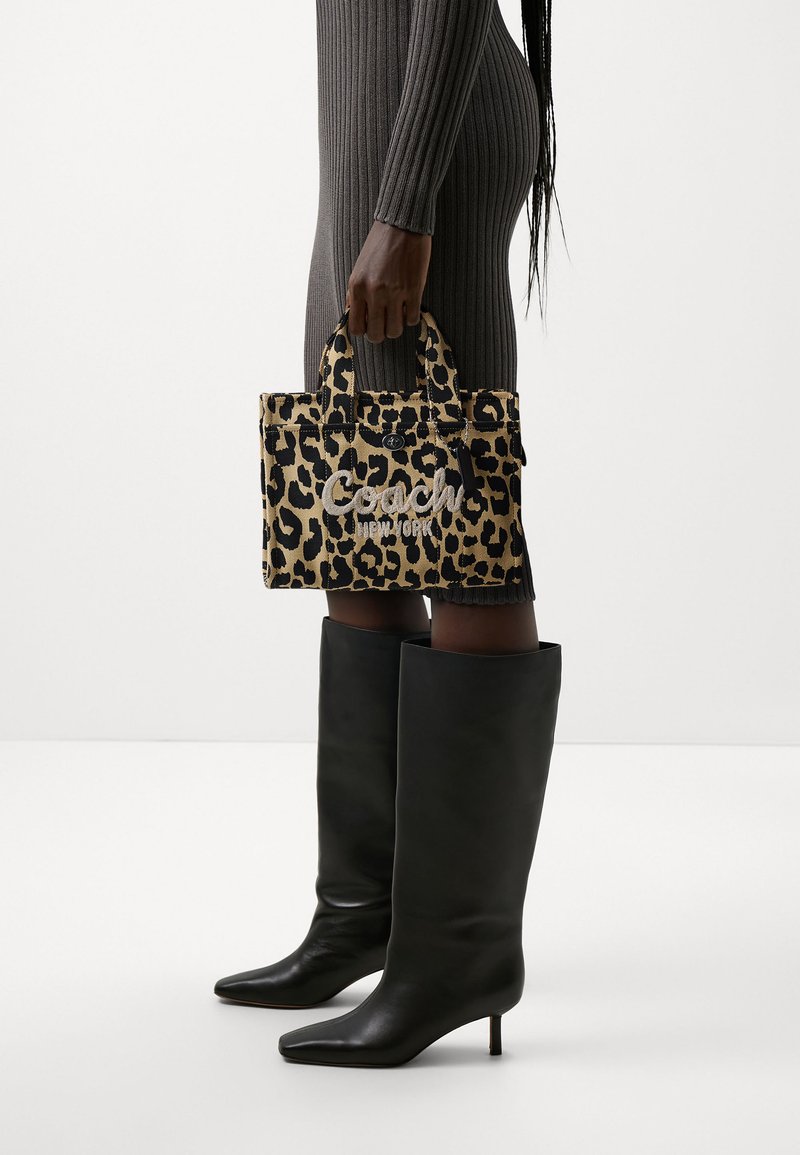 Sac à main à imprimé léopard avec le logo "Coach" brodé, doté de poignées doubles. Le modèle porte une robe moulante à côtes et des bottes noires hautes.