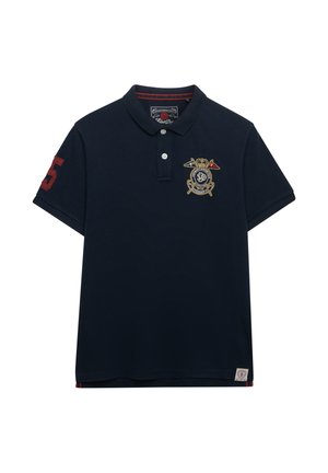 Marineblauw poloshirt met een rood nummer 5 applicatie op de mouw, een geborduurd wapen met gekruiste vlaggen op de borst, en twee witte knopen aan de hals.