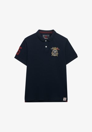 Marineblauw poloshirt met een rood nummer 5 applicatie op de mouw, een geborduurd wapen met gekruiste vlaggen op de borst, en twee witte knopen aan de hals.