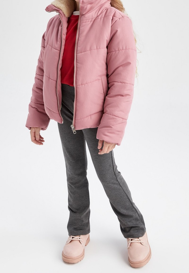 Enfant portant une veste matelassée rose avec col en peau retournée, chemise rouge, pantalon gris et bottes rose pâle, debout devant un fond blanc.