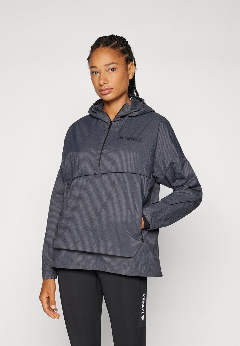 Giacca leggera con cappuccio in grigio scuro, realizzata in tessuto testurizzato, con mezza zip davanti e dettagli con logo sul petto. Abbinata a leggings neri.