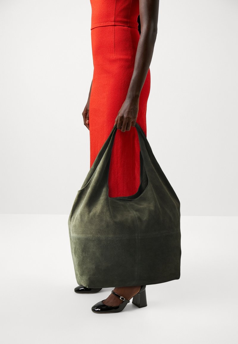 Personne en robe rouge tenant un grand sac cabas en daim vert foncé, portant des talons carrés en cuir verni noir avec des brides à la cheville.