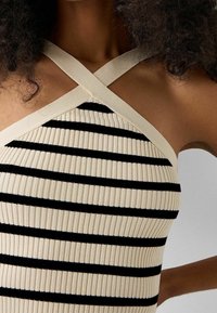 Débardeur côtelé de couleur crème avec des rayures horizontales noires. Il présente un col halter et des bretelles croisées.