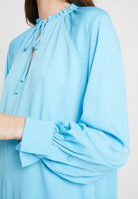 Blouse bleu clair avec manches longues bouffantes, poignets boutonnés et petit col à volants noué avec de fines cordelettes assorties.