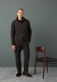 Giacca a zip marrone scuro, pantaloni affusolati abbinati e scarpe nere. Design semplice con una texture liscia. Sedia di legno sullo sfondo.